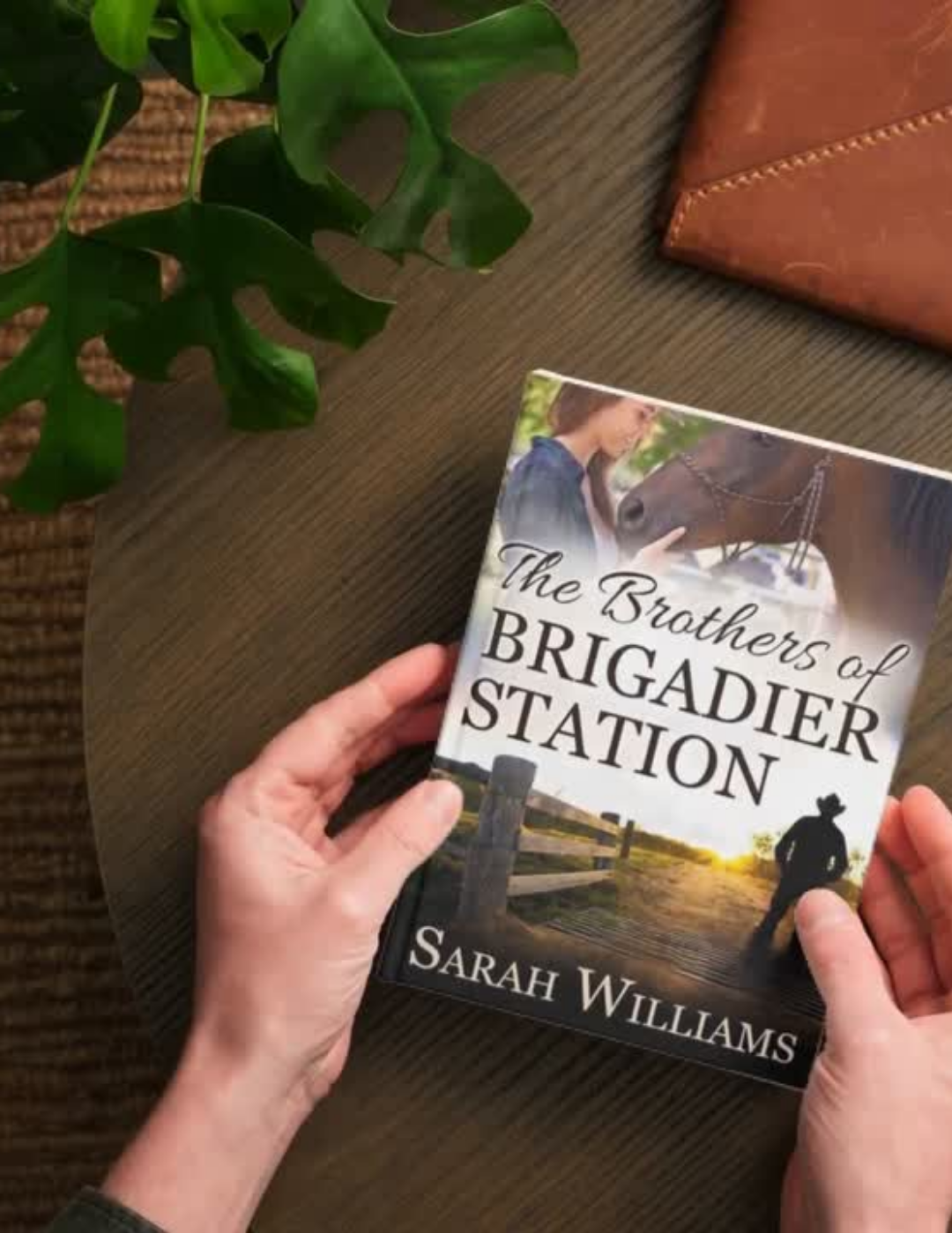Book titled 'The Brothers of Brigadier Station' by Sarah Williams held by hands on a wooden surface with a plant and leather item in the background.