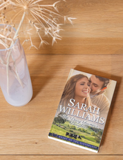 Book titled 'The Dairy Farmer's Daughter' by Sarah Williams on a wooden surface with a vase of flowers.