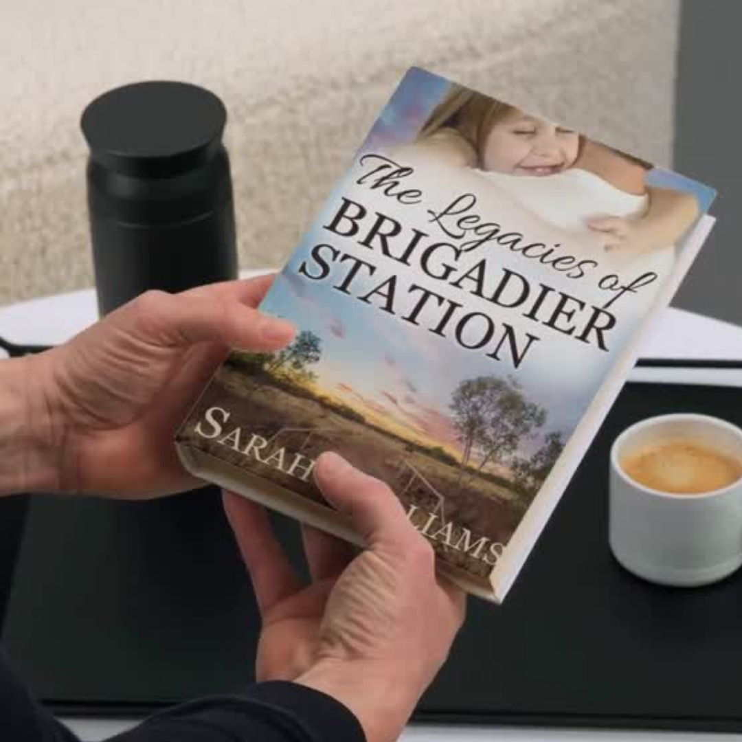 Person holding a book titled 'The Legacies of Brigadier Station' by Sarah Williams on a table with a cup of coffee.