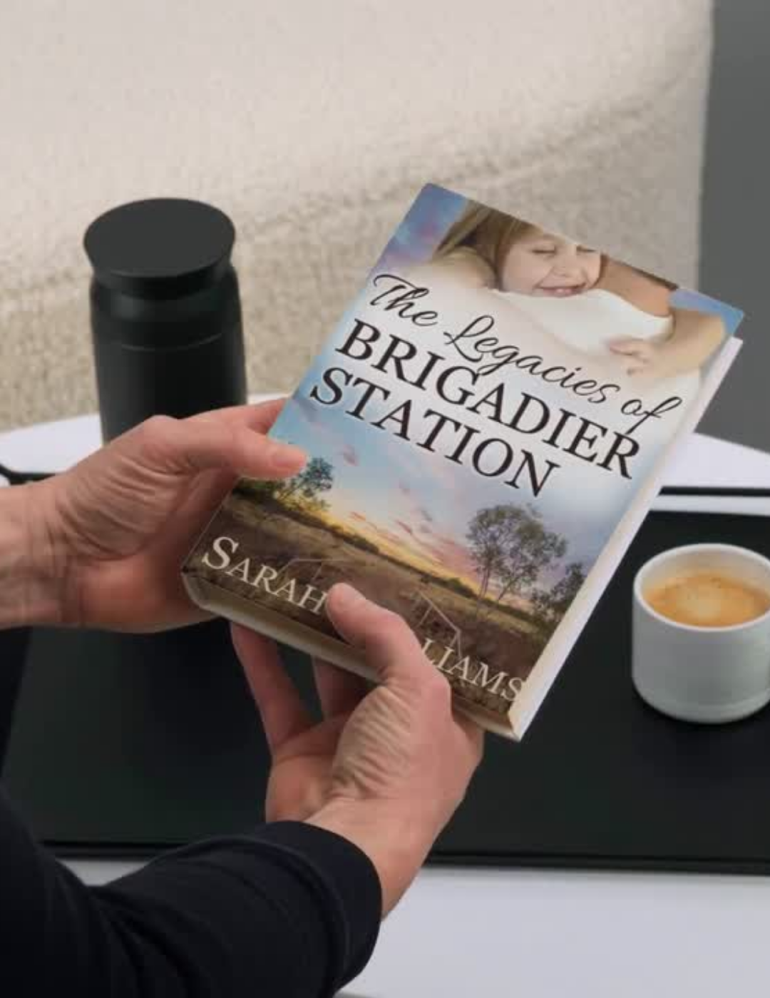 Person holding a book titled 'The Legacies of Brigadier Station' by Sarah Williams on a table with a cup of coffee.