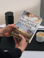 Person holding a book titled 'The Legacies of Brigadier Station' by Sarah Williams on a table with a cup of coffee.