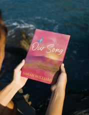 Person holding a book titled 'Our Song' by Sarah Williams with a scenic cover against a water background