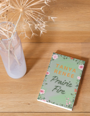 Book titled 'Prairie Fire' by Tanya Renee on a wooden surface with a vase of dried flowers.