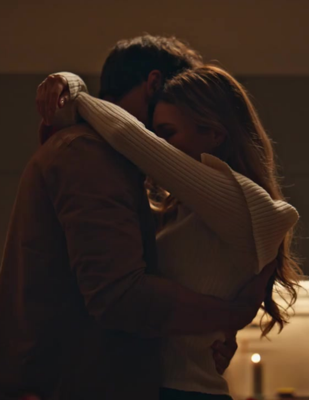 Two people embracing warmly in a dimly lit room with a candle in the background.