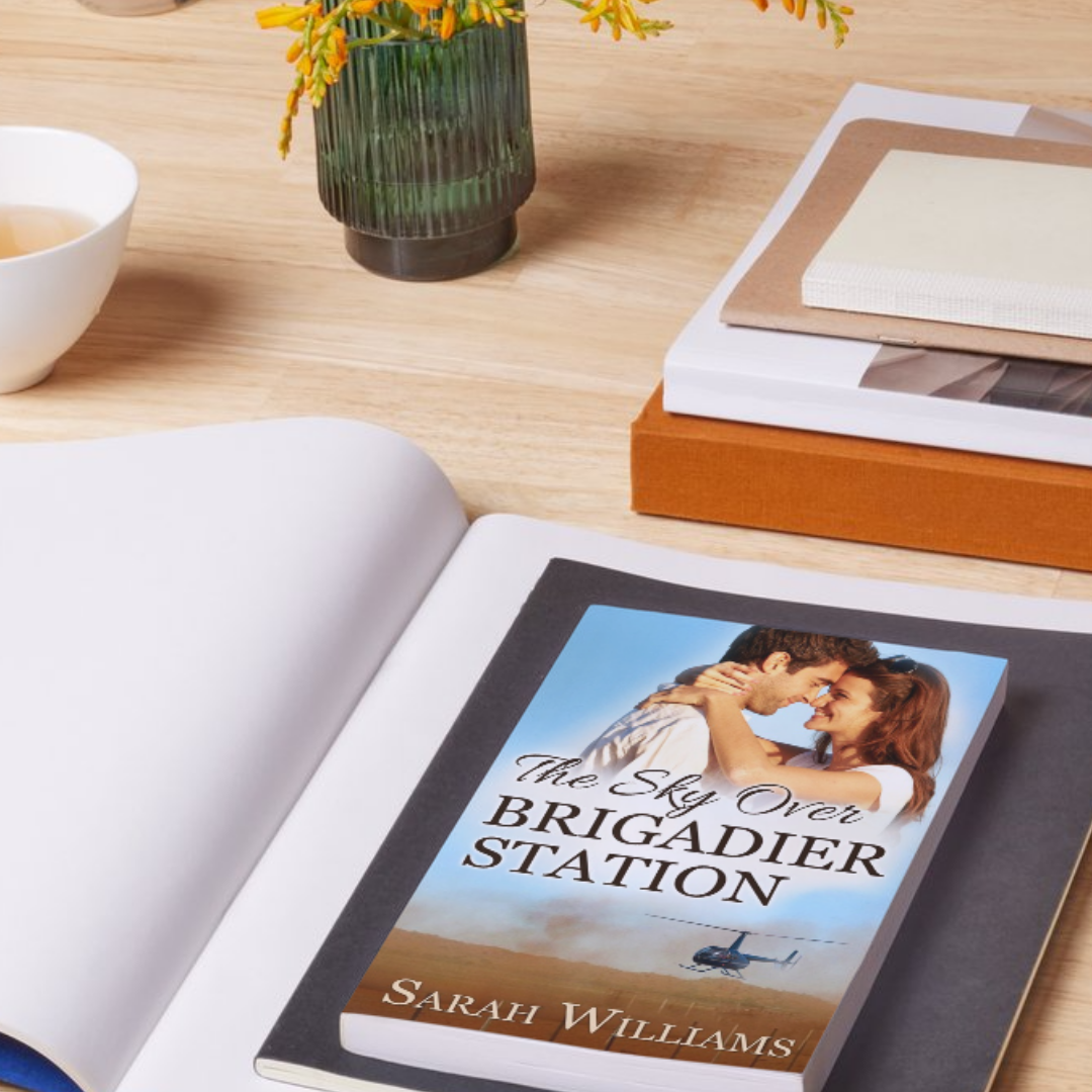 Book titled 'The Sky Over Brigadier Station' by Sarah Williams on a table with a vase of flowers and a cup.
