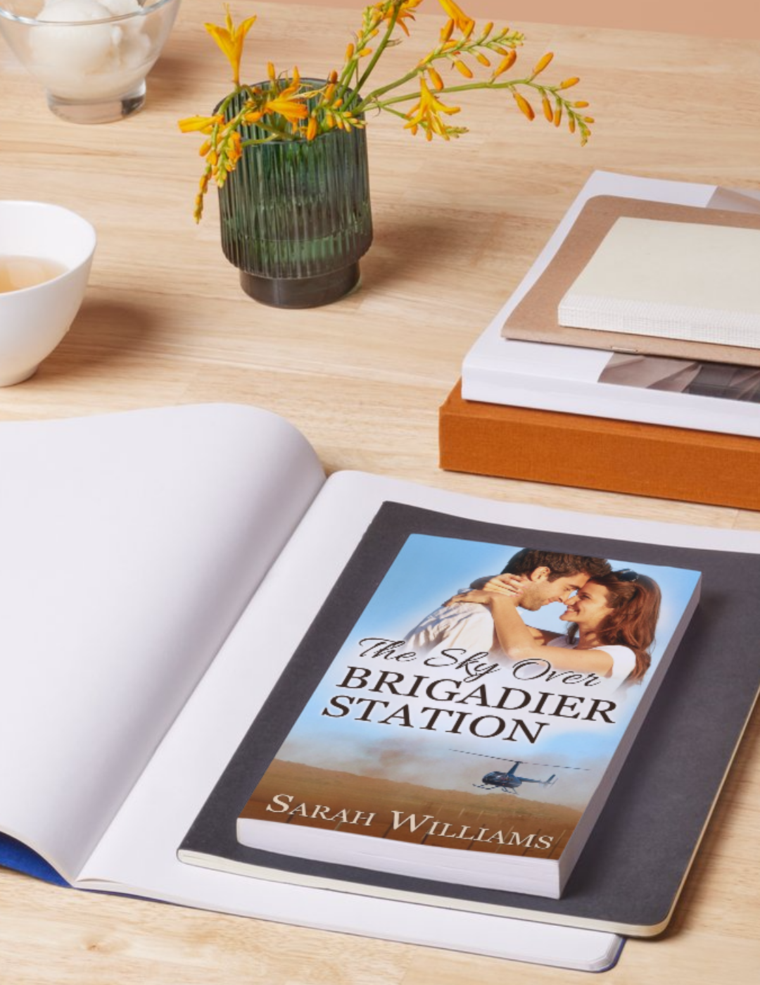 Book titled 'The Sky Over Brigadier Station' by Sarah Williams on a table with a vase of flowers and a cup.