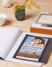 Book titled 'The Sky Over Brigadier Station' by Sarah Williams on a table with a vase of flowers and a cup.