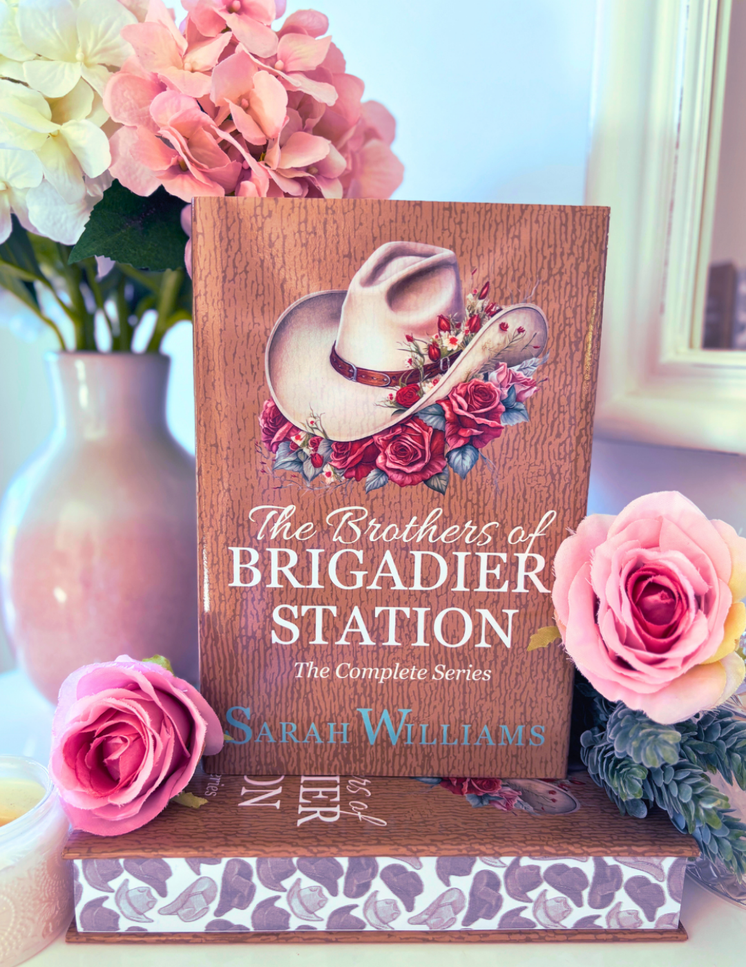 Book titled 'The Brothers of Brigadier Station' by Sarah Williams with floral design on a decorative stand, surrounded by flowers.
